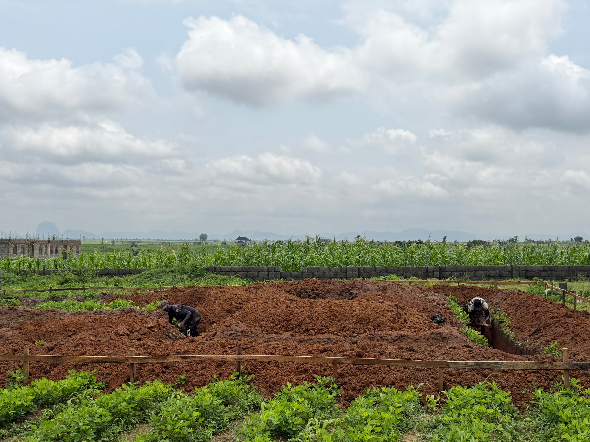 Foundation trenches being dug on a walled estate plot, farmland and hills behind, near Abuja
